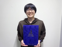 賞状入れを持って立つ茶色い服を着た学生