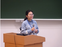 黒板の前に立つ水色のセーターの女性